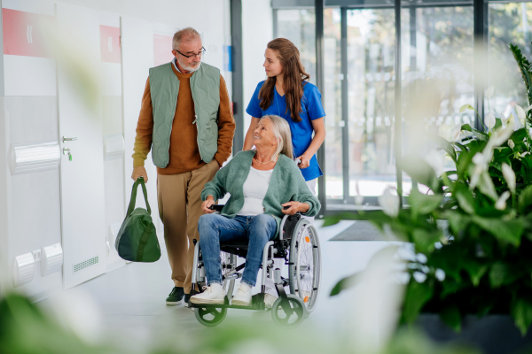 Young nurse pushing wheelchair and talking with senior patients at hospital corridor.