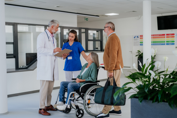 Mature doctor and his young colleaguetalking with his senior patients at hospital corridor.