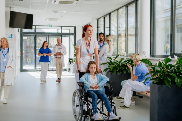 Young nurse pushing little girl on wheelchair at a hospital corridor.