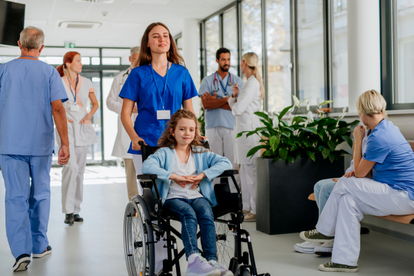 Young nurse pushing little girl on wheelchair at a hospital corridor.