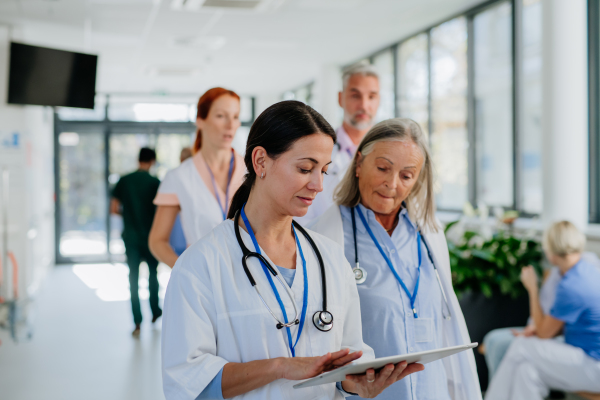 Older doctor giving advise to her younger colleague, discussing at a hospital corridor. Health care concept.