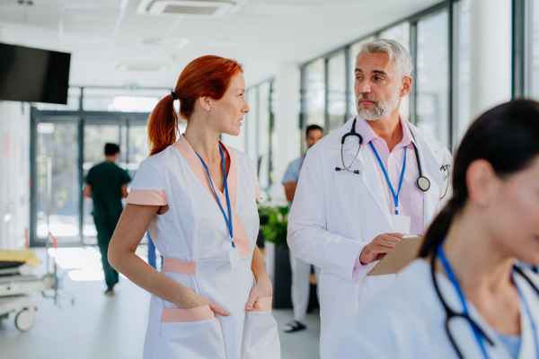 Older doctor giving advise to his younger colleague, discussing at a hospital corridor. Health care concept.