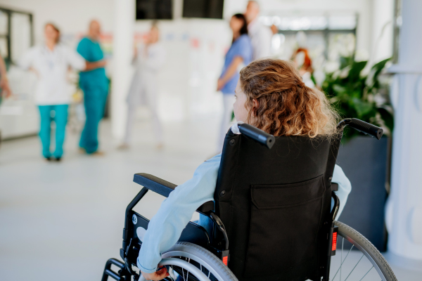 Rear view of little girl with teddy bear sitting on a wheelchair.