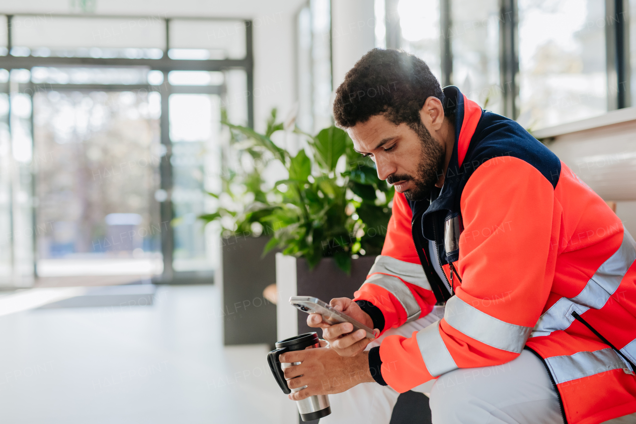 Rescuer having break, drinking coffee and scrolling his mobile phone at a hospital corridor.