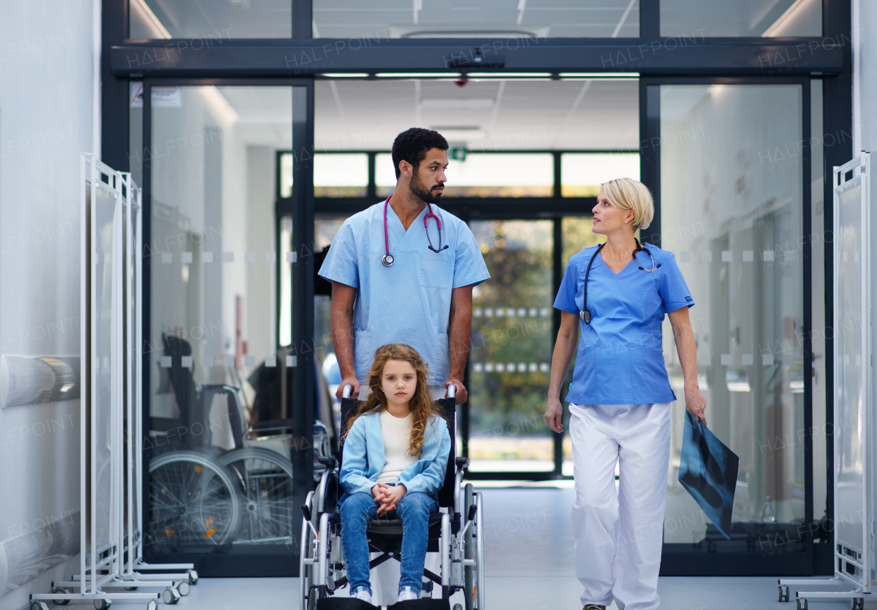 Pregnant doctor with her colleague pushing litle girl at wheelchair at the pediatric department.