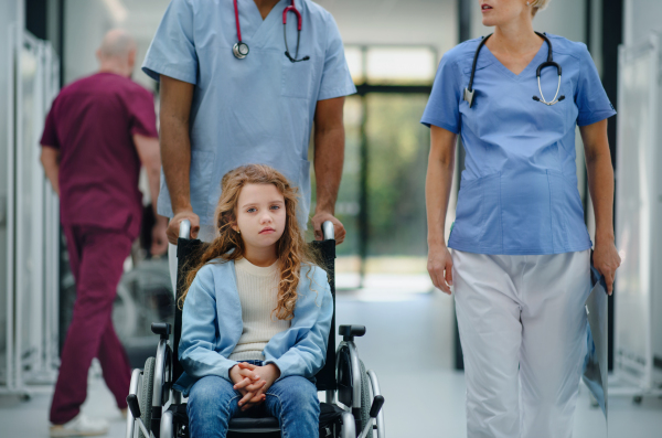 Pregnant doctor with her colleague pushing litle girl at wheelchair at the pediatric department.