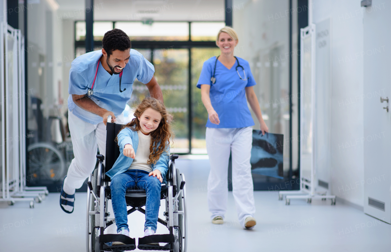 Young doctor pushing little girl at wheelchair at the pediatric corridor, having fun.