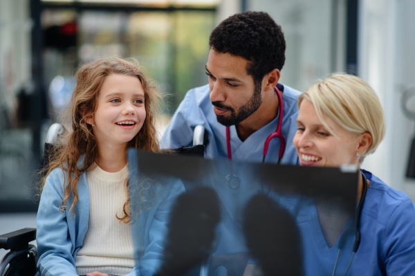 Young multiracial doctor with his colleague showing x-ray image of lungs to little patient on wheelchair.