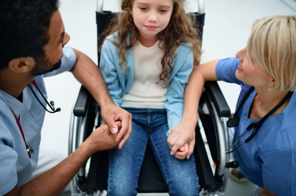 Young multiracial doctor with his colleague consoling little girl on a wheelchair.