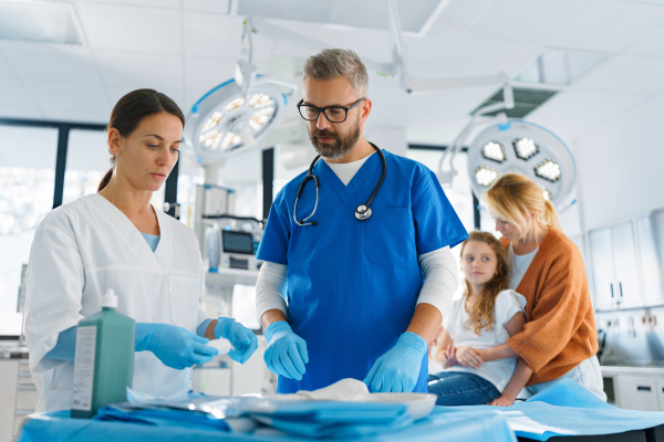 Mature doctor with nurse preparing examinating tools, little girl with mother sitting in the background.