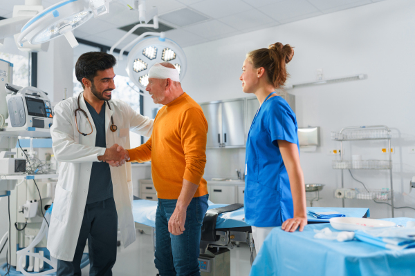 Senior man with injured head leaving surgery, shaking hand with young multiracial doctor.