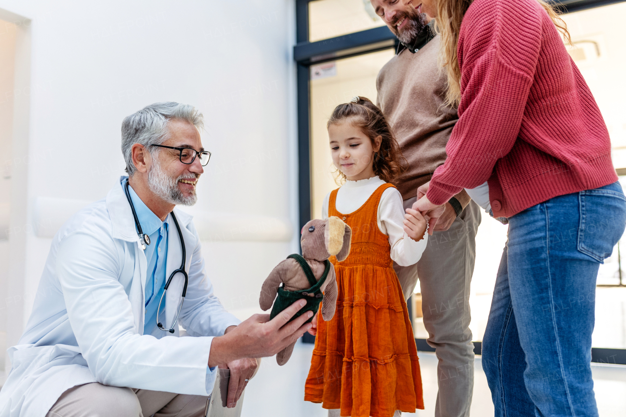 Pediatrician looking at girl's toy. Girl patient is at examination in a modern clinic with her parents. Emotional support during medical examination in hospital.