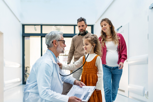 Girl listening pediatrician heartbeat with stethoscope. Girl patient is at examination in a modern clinic with her parents. Emotional support during medical examination.