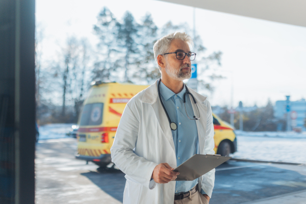 Portrait of mature doctor in hospital corridor. Handsome doctor with gray hair wearing white coat, stethoscope around neck standing in modern private clinic.