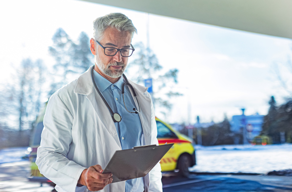 Portrait of mature doctor in hospital corridor. Handsome doctor with gray hair wearing white coat, stethoscope around neck standing in modern private clinic.