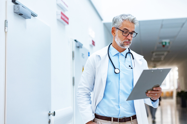 Portrait of mature doctor in hospital corridor. Handsome doctor with gray hair wearing white coat, stethoscope around neck standing in modern private clinic.