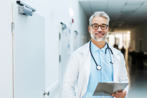 Portrait of smiling doctor standing in hospital corridor. Handsome doctor with gray hair wearing white coat, stethoscope around neck standing in modern private clinic and looking at camera.