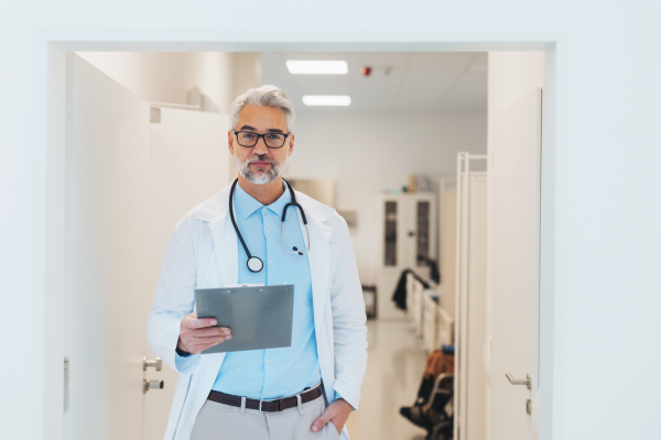 Portrait of mature doctor in hospital corridor. Handsome doctor with gray hair wearing white coat, stethoscope around neck standing in modern private clinic.