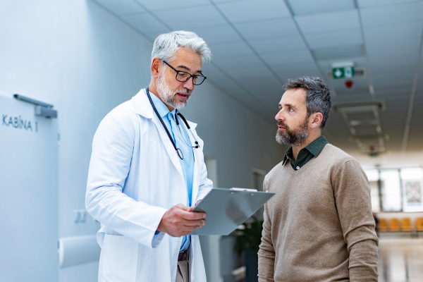 Doctor talking with patient in hospital corridor. Handsome doctor with gray hair holding clipboard with test result, consulting with man, standing in modern private clinic.