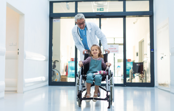 Friendly pediatrician pushing little patient in wheelchair. Cute preschool girl trusting her healthcare workers in hospital. Concept of children healthcare and emotional support for child patients.