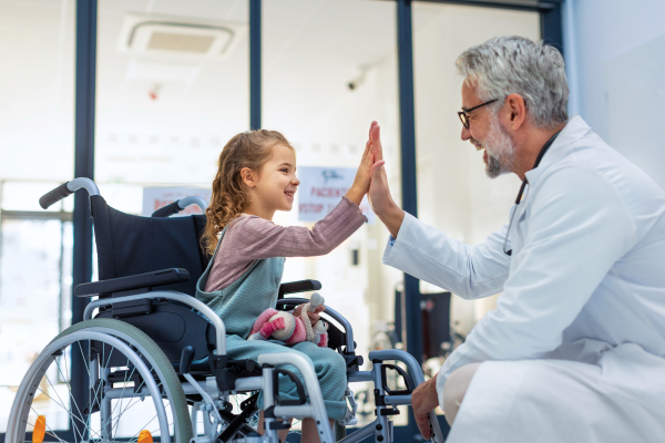 Friendly pediatrician giving high five to little patient in wheelchair. Cute preschool girl in wheelchair greeting doctor in hospital. Concept of children healthcare and emotional support for child patients.