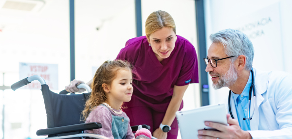 Friendly pediatrician showing someting on tablet to little patient in wheelchair. Cute preschool girl trusting her doctor and nurse in hospital. Concept of children healthcare and emotional support for child patients.