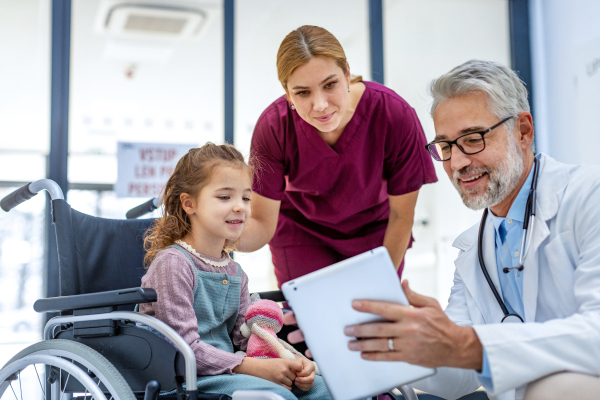 Friendly pediatrician showing someting on tablet to little patient in wheelchair. Cute preschool girl trusting her doctor and nurse in hospital. Concept of children healthcare and emotional support for child patients.