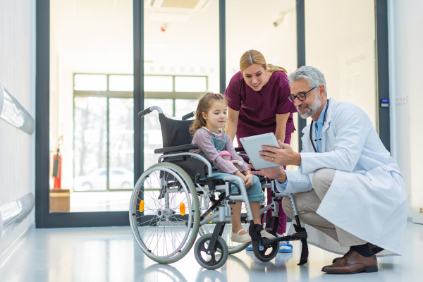 Friendly pediatrician showing someting on tablet to little patient in wheelchair. Cute preschool girl trusting her doctor and nurse in hospital. Concept of children healthcare and emotional support for child patients.