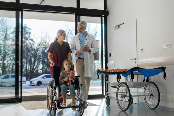 Friendly pediatrician nurse talking, pushing little patient in wheelchair. Cute preschool girl trusting her doctor and nurse in hospital. Concept of children healthcare and emotional support for child patients.