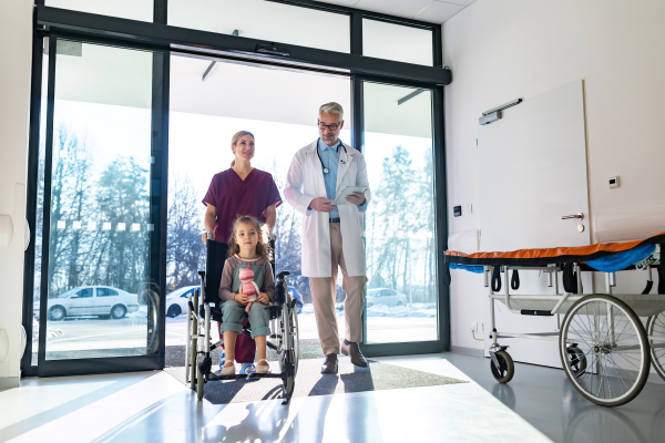 Friendly pediatrician walking with little patient in wheelchair. Nurse pushing wheelchair. Cute preschool girl trusting her doctor and nurse in hospital. Concept of children healthcare and emotional support for child patients.