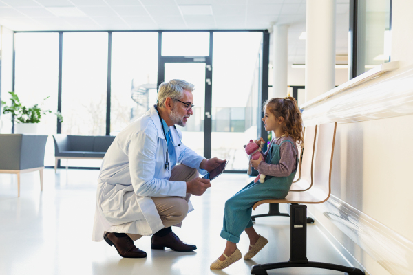 Friendly pediatrician talking to little patient. Cute preschool girl with doctor in hospital corridor. Concept of children healthcare and emotional support for child patients.