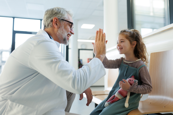 Friendly pediatrician giving high five to little patient. Cute preschool girl in greeting doctor in hospital corridor.Concept of children healthcare and emotional support for child patients.