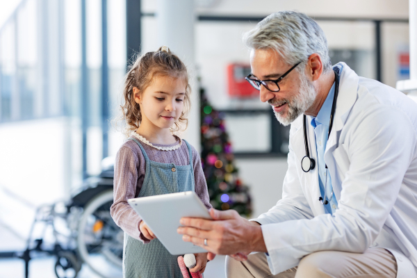 Friendly pediatrician showing someting on tablet to little patient. Cute preschool girl trusting her doctor talking with him. Concept of children healthcare and emotional support for child patients.