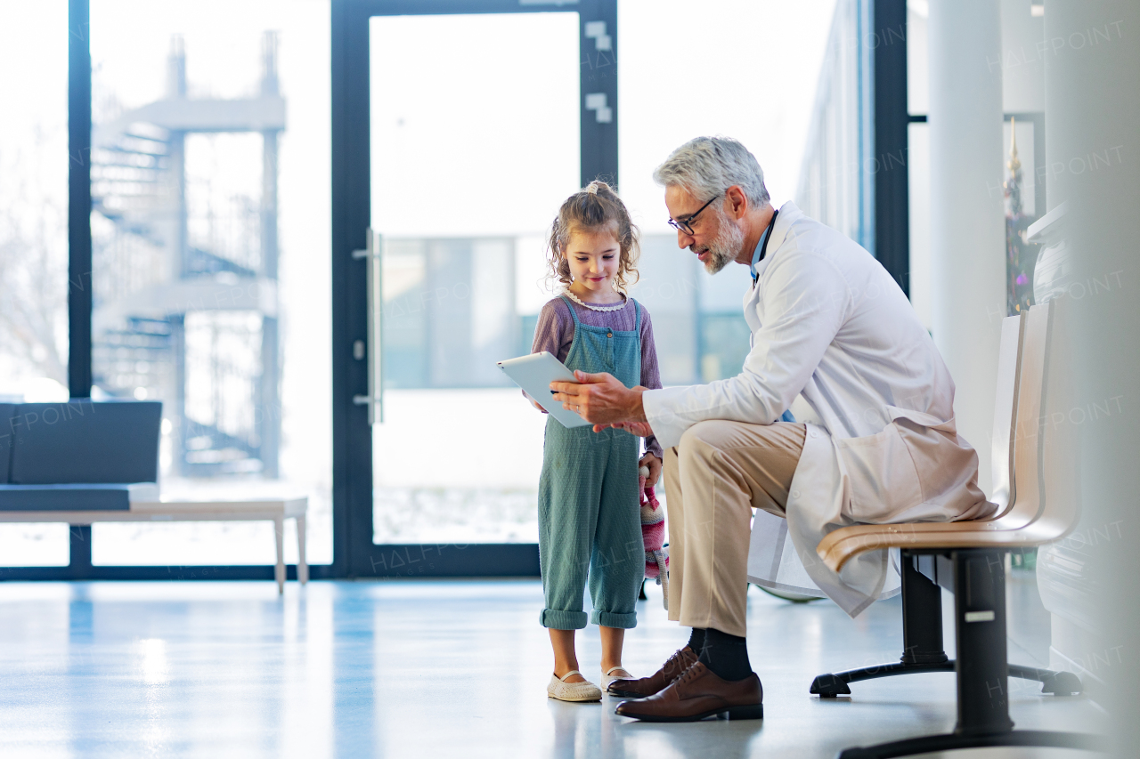 Friendly pediatrician showing someting on tablet to little patient. Cute preschool girl trusting her doctor talking with him. Concept of children healthcare and emotional support for child patients.