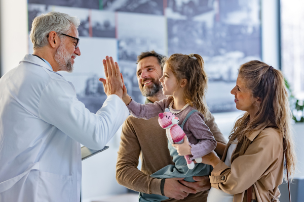 Friendly pediatrician giving high five to little patient. Parents with girl patient greeting doctor in hospital corridor. Concept of children healthcare and emotional support for child patients.