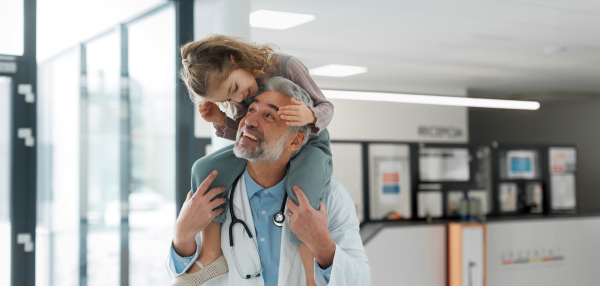 Portrait of a pediatrician with a little patient sitting on his shoulders, piggyback. Doctor carrying girl on shoulders. Friendly relationship between the doctor and the child patient.