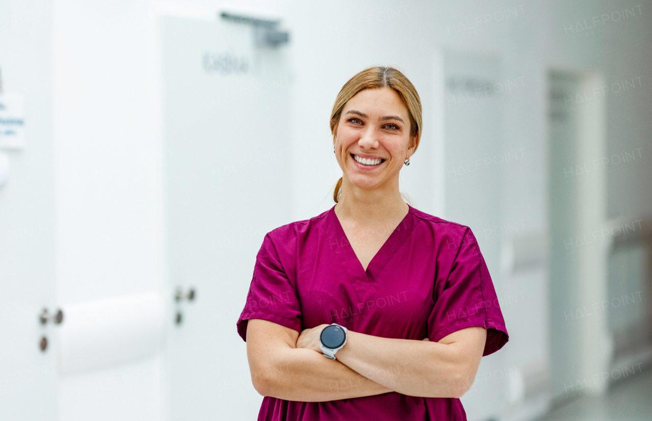 Portrait of a beautiful nurse in uniform, standing in the hospital corridor with her arms crossed, looking at the camera and smiling.