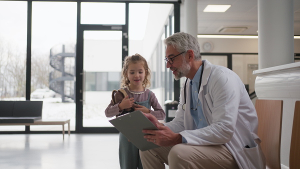 Friendly pediatrician talking to little patient. Cute preschool girl with doctor in hospital corridor. Concept of children healthcare and emotional support for child patients.