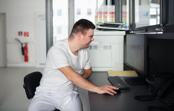 Young man with down syndrome working in a hospital office, writing something on computer. Concept of integration people with disabilities into society.