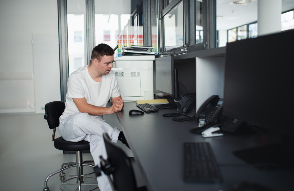 Young man with down syndrome working in a hospital office, writing something on computer. Concept of integration people with disabilities into society.