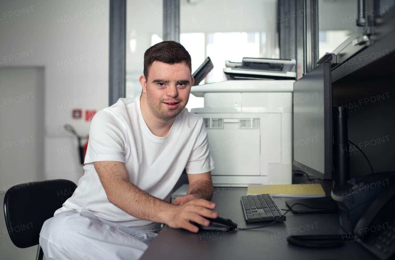 Young man with down syndrome working in a hospital office, writing something on computer. Concept of integration people with disabilities into society.