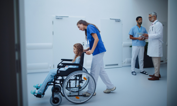 Young nurse pushing little girl on wheelchair at a hospital corridor.