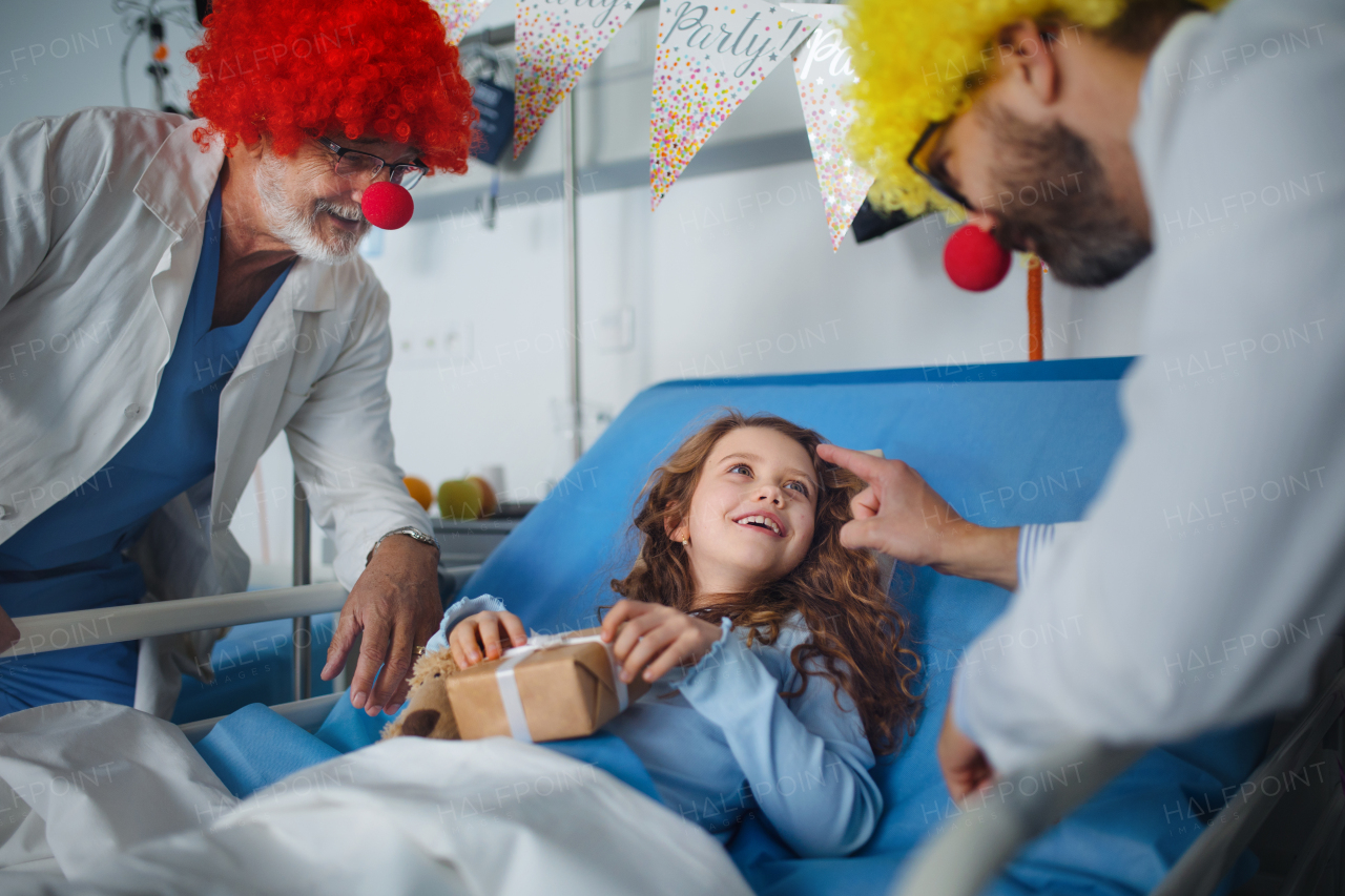 Happy doctors with clown red noses celebrating birthday with little girl in a hospital room.