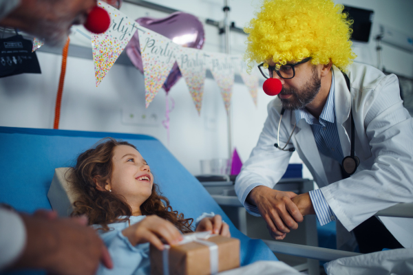 Happy doctor with clown red noses celebrating birthday with little girl in a hospital room.