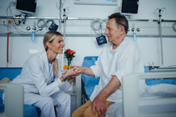 Young woman doctor holding hand, and talking with her patient in hospital room.