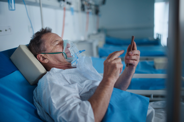 Senior man with oxygen mask lying in the hospital bed and scrolling smartphone.