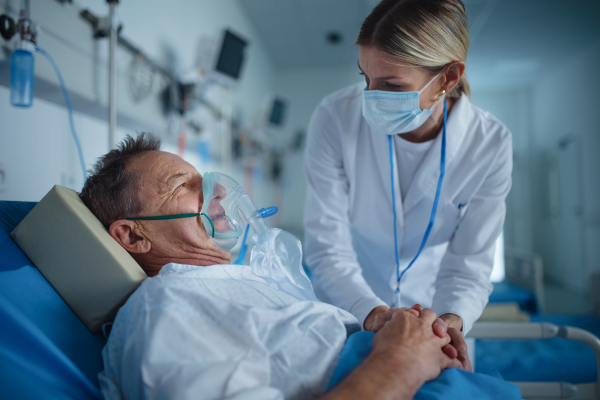 Young woman doctor talking to elderly patient with oxygen mask in hospital room.