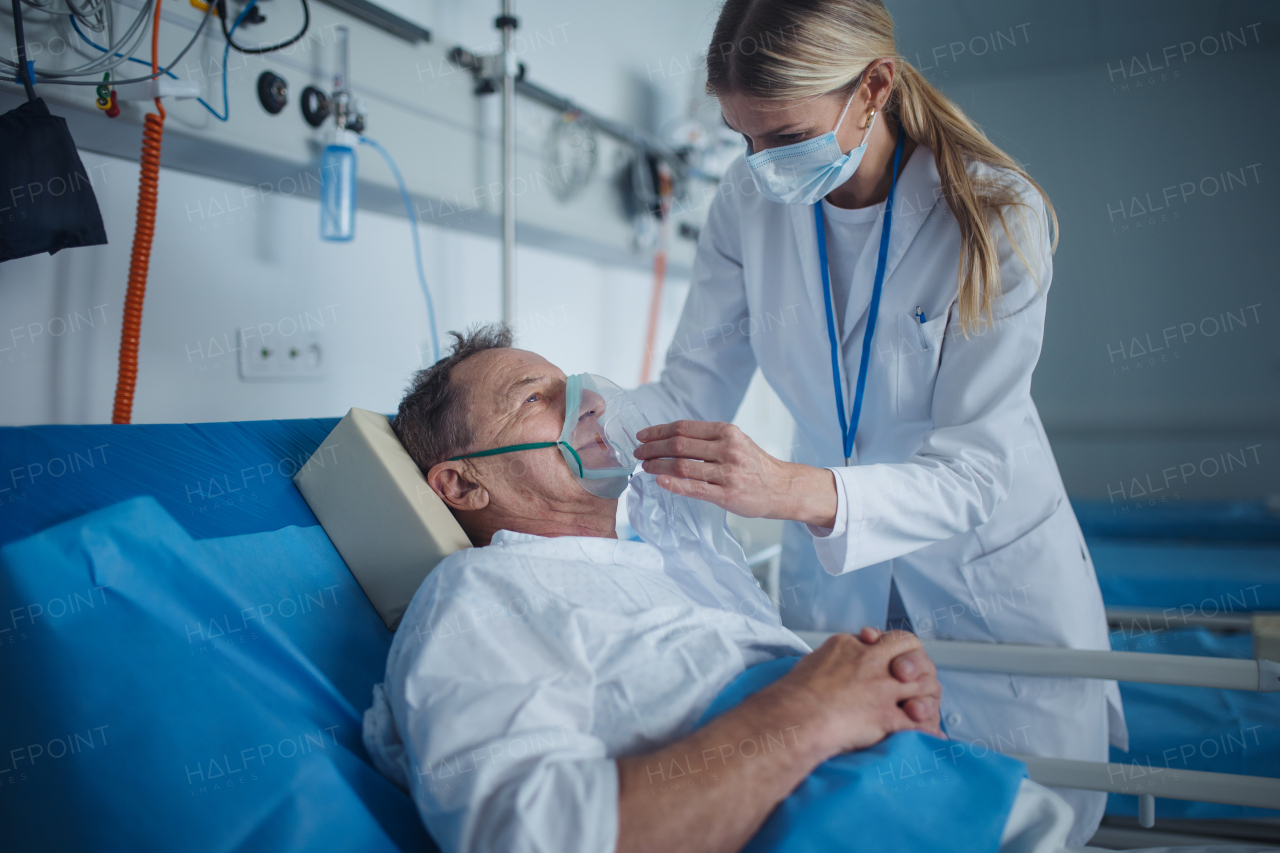 Young woman doctor putting on oxygen mask to elderly patient lying in hospital room.