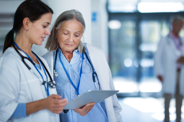 Older doctor giving advise to his younger colleague, discussing at a hospital corridor. Health care concept.