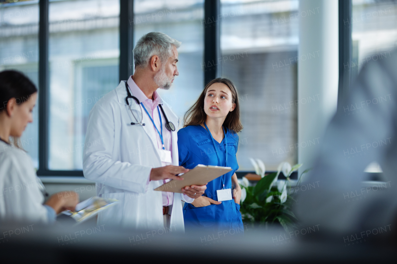 Older doctor giving advise to his younger colleague, discussing at a hospital corridor. Health care concept.